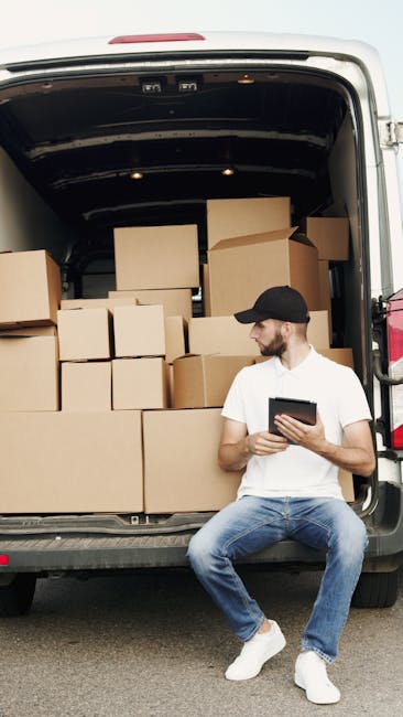 A man wearing a white t-shirt, blue jeans, white sneakers, and a black cap is sitting on the pavement beside the open rear doors of a van used for home relocation. The van's cargo area is filled with an assortment of packed cardboard boxes, some of which are large, while others are smaller, all secured with packing tape. The boxes are arranged in a layered manner, with a few stacked directly on top of each other, and some positioned against the van's interior walls, indicating an organized loading process typical of furniture transport and packing for a move. The man is holding a tablet, possibly coordinating the packing or move details, and appears to be part of a professional removals service, such as Man With a Van Ladbroke Grove. The background suggests an outdoor setting, with natural light illuminating the scene, capturing the essential elements involved in a house removal, from packing to loading logistics.
