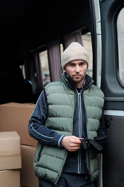 A man dressed in a light grey knitted beanie, a dark blue jacket with white piping, and a green padded vest stands inside the cargo area of a van used for home relocation. Behind him, several cardboard moving boxes of various sizes are arranged on a flat surface, ready for packing and transportation. The interior of the van is dimly lit, with dark upholstery visible in the background. The man is holding a black strap, likely to secure furniture or boxes during the loading process, which is part of the furniture transport and packing and moving tasks carried out by Man With a Van Ladbroke Grove. The scene depicts the loading phase of a house removal, with the van partially open providing access to the interior, situated on a paved area, preparing for a professional relocation service within W10, Ladbroke Grove.