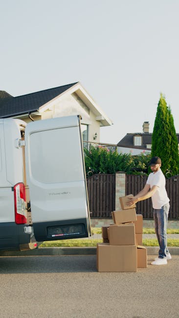 A man wearing a white t-shirt, blue jeans, white sneakers, and a black cap is sitting on the pavement beside the open rear doors of a van used for home relocation. The van's cargo area is filled with an assortment of packed cardboard boxes, some of which are large, while others are smaller, all secured with packing tape. The boxes are arranged in a layered manner, with a few stacked directly on top of each other, and some positioned against the van's interior walls, indicating an organized loading process typical of furniture transport and packing for a move. The man is holding a tablet, possibly coordinating the packing or move details, and appears to be part of a professional removals service, such as Man With a Van Ladbroke Grove. The background suggests an outdoor setting, with natural light illuminating the scene, capturing the essential elements involved in a house removal, from packing to loading logistics.