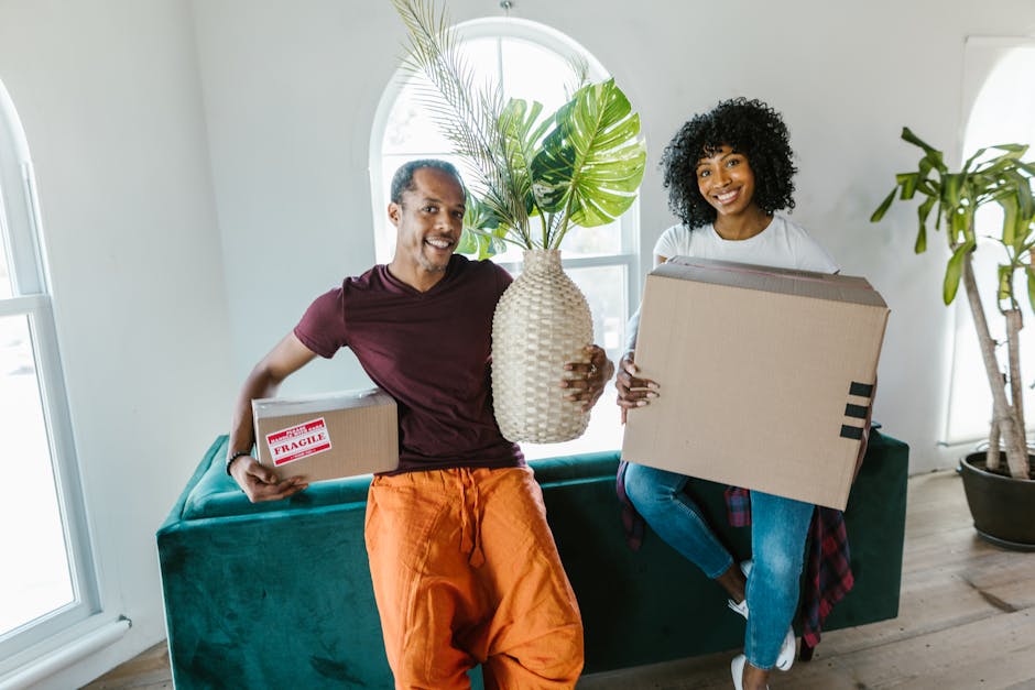 A man and a woman inside a bright, airy room with large arched windows, engaged in a home relocation process managed by Man With a Van Ladbroke Grove. The man, wearing a maroon t-shirt and orange trousers, is holding a cardboard box labeled 'FRAGILE' in one hand and a large ceramic vase with broad green palm leaves in the other. The woman, dressed in a white top and blue jeans, is sitting on a teal sofa and carrying a medium-sized cardboard box. The scene includes natural daylight illuminating the space, with visible packaging materials such as cardboard boxes and protective coverings, indicating an active packing and moving process. In the background, there's a tall potted plant and the minimal modern decor of the room, suggesting a recent or ongoing home transfer, with furniture and packed items prepared for transport by professional removals services.