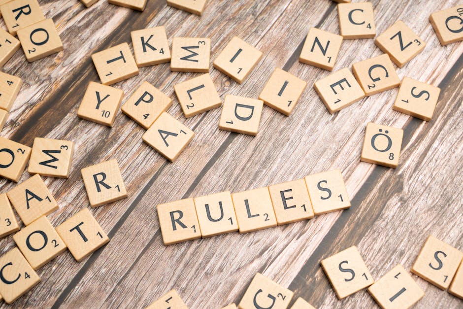 Photograph of numerous wooden Scrabble tiles scattered across a wooden surface, with some tiles forming the word 'RULES' in the center. Other tiles display letters and numbers, such as 'A', 'Y', 'OM',
