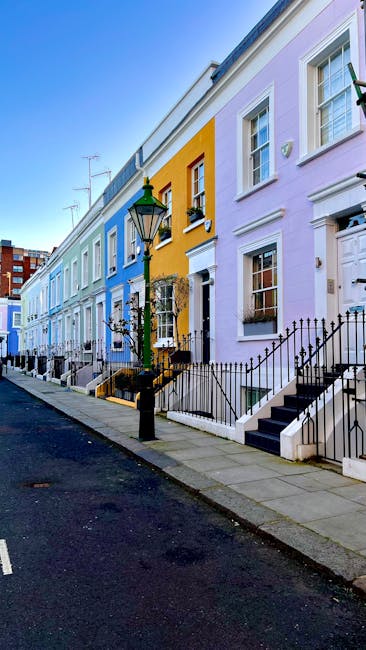 A row of colorful terraced houses on Ladbroke Grove, with pastel facades in shades of blue, yellow, pink, and purple, each featuring white-framed windows, black iron railings, and external stairs leading to front doors. The pavement is made of concrete slabs, with a classic black lamp post situated on the sidewalk near the middle of the image. The street appears quiet with no visible vehicles or pedestrians, and the sky is clear and bright. The scene reflects a typical residential area suitable for home relocation or furniture transport purposes, with the houses’ external appearance indicating a period or Victorian style. Man With a Van Ladbroke Grove specializes in removals and moving services, serving areas like Kensington and Chelsea, where such streets are common for house removals and furniture transport. The image emphasizes the professional process of planning a house move within this vibrant neighborhood, with no moving activity in progress visible at the moment.