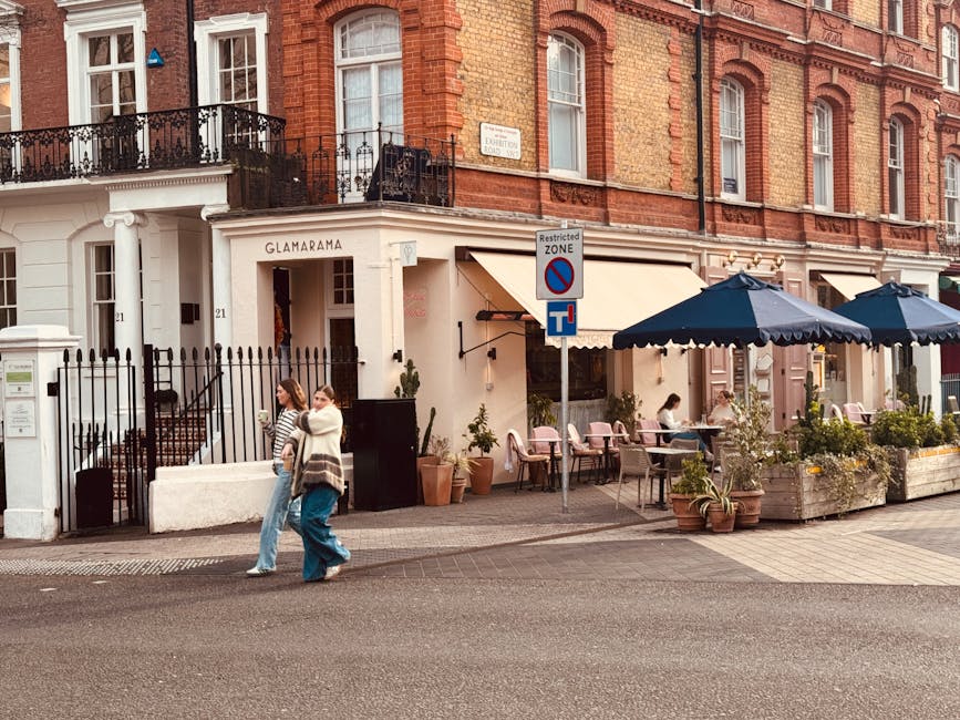 A row of colorful terraced houses on Ladbroke Grove, with pastel facades in shades of blue, yellow, pink, and purple, each featuring white-framed windows, black iron railings, and external stairs leading to front doors. The pavement is made of concrete slabs, with a classic black lamp post situated on the sidewalk near the middle of the image. The street appears quiet with no visible vehicles or pedestrians, and the sky is clear and bright. The scene reflects a typical residential area suitable for home relocation or furniture transport purposes, with the houses’ external appearance indicating a period or Victorian style. Man With a Van Ladbroke Grove specializes in removals and moving services, serving areas like Kensington and Chelsea, where such streets are common for house removals and furniture transport. The image emphasizes the professional process of planning a house move within this vibrant neighborhood, with no moving activity in progress visible at the moment.