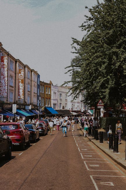 A busy street scene in Ladbroke Grove showcasing a line of parked cars along the curb and a sidewalk filled with pedestrians, some carrying shopping bags, with several storefronts featuring blue awnings and colorful facades visible in the background. Tall trees with lush green foliage shade parts of the street, and a large leafy tree extends over the sidewalk on the right. The road appears to be temporarily blocked off for pedestrian use or a community event, with bollards and traffic signs marking the area. This setting reflects a vibrant urban environment typical of residential and commercial areas in Ladbroke Grove, often associated with home relocation and moving services. The scene captures the lively atmosphere of a populated neighbourhood, relevant to house removals and furniture transport activities carried out by companies like Man With a Van Ladbroke Grove, as they facilitate packing, loading, and moving household items within this bustling community.