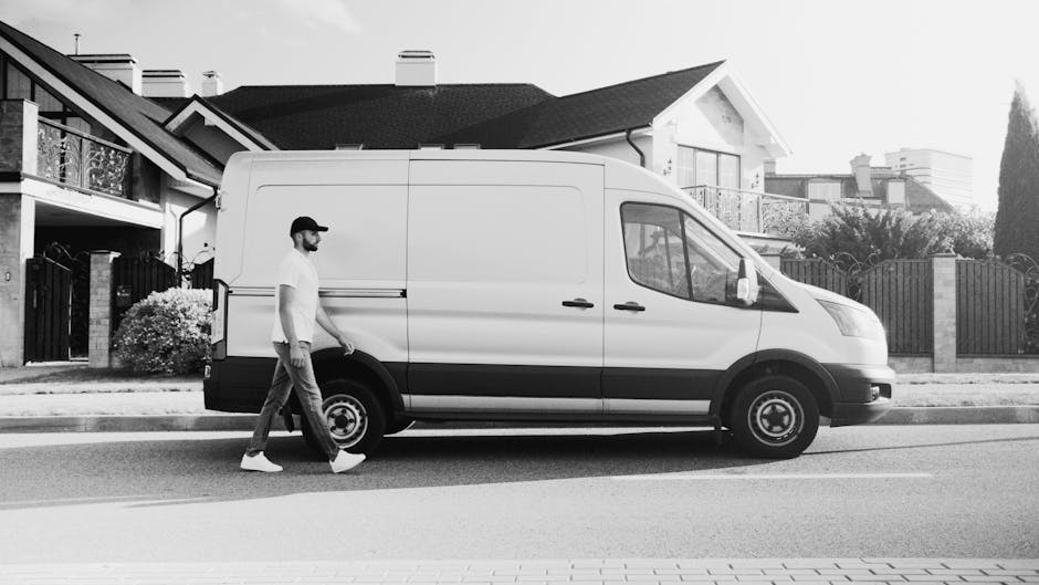 A man with a beard, wearing a baseball cap, white t-shirt, and jeans, walks beside a white panel van parked on the street in a residential neighbourhood. The van is positioned parallel to the pavement, with its sliding side door closed and the front facing right. In the background, there are modern houses with tiled roofs, balconies, and fencing, along with some trees and shrubs. The scene appears to be part of a home relocation or furniture transport process, with the man possibly preparing for a moving or loading activity as part of a professional removals service provided by Man With a Van Ladbroke Grove. The surroundings are well-lit, indicating daytime, and the pavement is clean with visible road markings for parking or stopping zones.