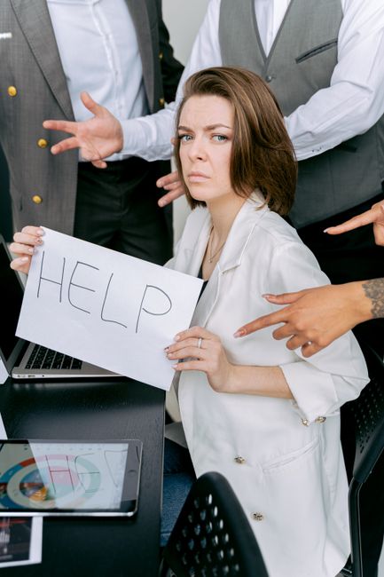 A woman with shoulder-length brown hair and a concerned expression is holding a white sign with the word 'HELP' written in large black letters. She is seated at a desk with a laptop and a tablet visible in front of her. Surrounding her are several individuals, some pointing and gesturing towards her, indicating a tense or urgent situation. The setting appears to be an office environment, with professional attire and a group engaged in a serious discussion related to a work or business crisis. This image highlights themes of distress, communication, and the need for assistance, relevant to professional client support or crisis management services. The context aligns with providing help in challenging scenarios, similar to the logistical or emotional challenges encountered during house removals or relocation processes.