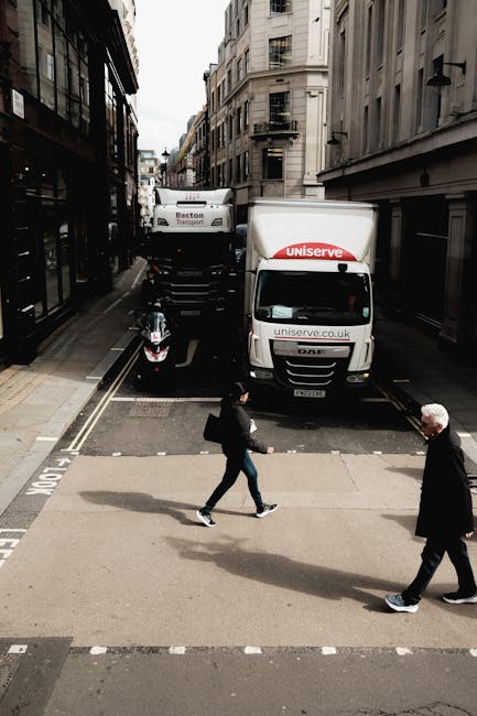 A street scene in an urban area shows a man with a black jacket and backpack crossing the road while an older man in a dark coat walks in the opposite direction. Behind them, a white delivery van branded with 'Uniserve' and a black lorry with 'Becton Transport' are parked on the road, partially blocking the street. The vehicles are positioned near a loading area adjacent to tall, multi-storey buildings with classic architectural features and fire escapes. A motorcycle is parked close to the curb near the van. The scene is captured during daylight with natural light illuminating the surroundings, indicating preparation or transition related to home relocation or furniture transport services, with Man With a Van Ladbroke Grove offering assistance in such moving logistics.