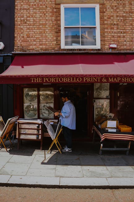 A person dressed in a light blue shirt and dark trousers is standing outside a brick building on Portobello Road, reading a menu or signboard. The storefront has a red retractable awning with gold lettering that reads 'The Portobello Print & Map Shop.' In front of the shop, there are wooden and fabric-upholstered chairs with striped cushions, along with various cardboard boxes and display materials arranged on the pavement. The scene is illuminated by daylight, and an upstairs window with a white frame is visible above the store, reflecting the blue sky and some surrounding buildings. This setting captures a typical street scene of a shop involved in a home relocation or packing and moving process, aligning with house removals and furniture transport services offered by Man With a Van Ladbroke Grove.