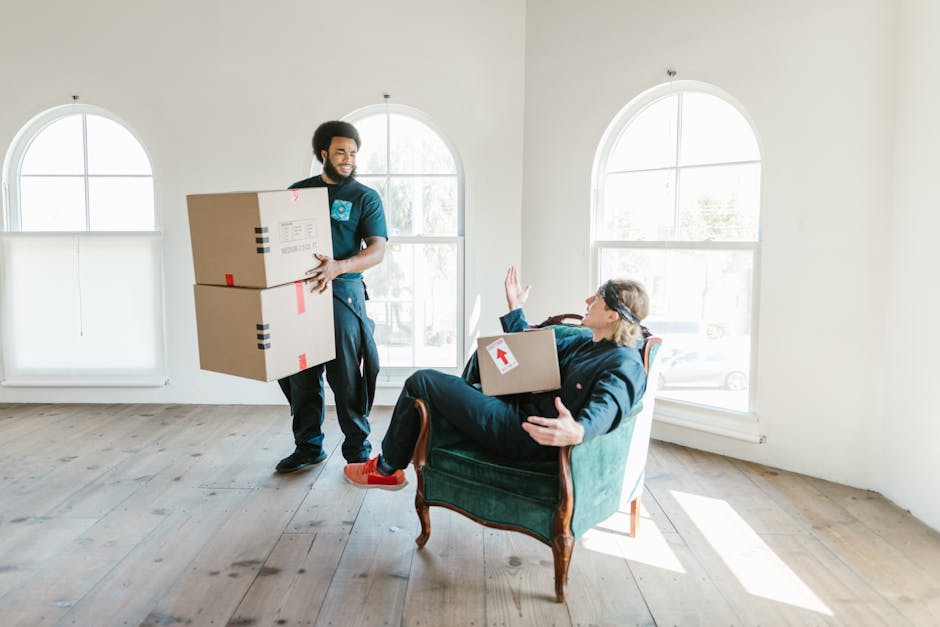 A professional removal worker from Man With a Van Ladbroke Grove is inside a bright and spacious room with large arched windows, carrying two cardboard boxes secured with red and black packing tape. The worker is dressed in a blue uniform and is smiling while holding the boxes, indicating careful handling during the home relocation process. Inside the room, a woman seated in a dark blue upholstered armchair is visible, holding a smaller box with an upward-pointing arrow label and raising her hand as if greeting or signalling completion. The room has light-colored wooden flooring and minimal decor, with natural sunlight illuminating the space, suggesting a well-organized packing and moving operation as part of the furniture transport and packing process managed by [COMPANY_NAME]. The scene captures the logistics involved in house removals, highlighting the careful packing, loading, and coordination typical of professional move services in the Portobello Road area.