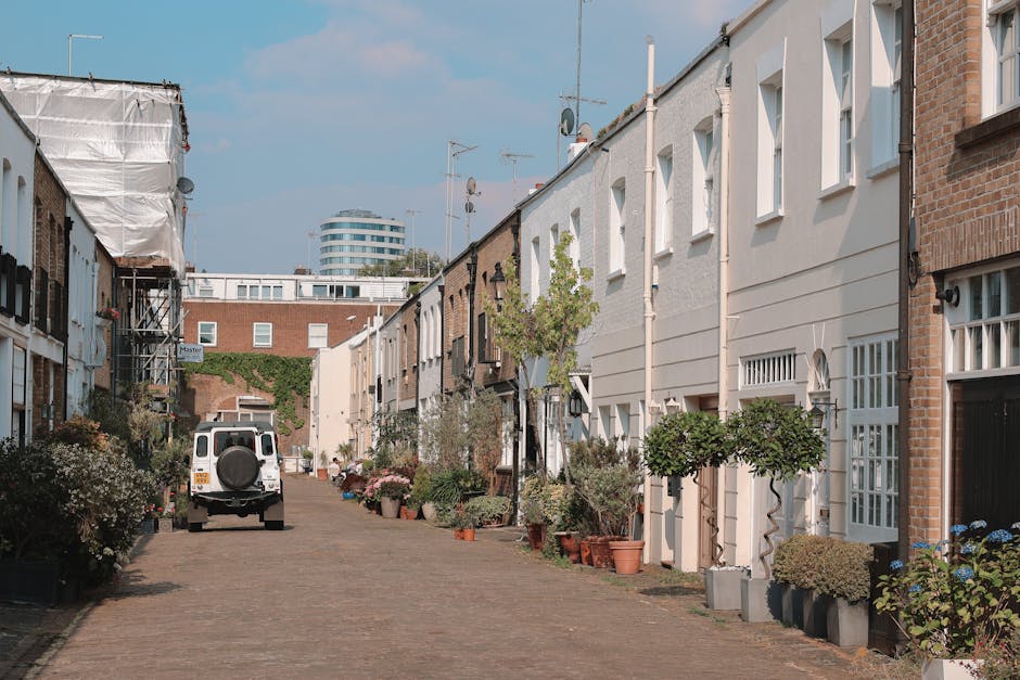A narrow residential street in Golborne Road, Ladbroke Grove, with a row of terraced houses on each side. The houses are a mix of white and brick facades, with some featuring small front gardens filled with potted plants and trees. The street is paved with cobblestones and has a white van parked near the center, likely used for a home relocation or furniture transport. Several large cardboard boxes and wrapped pieces of furniture are visible, being loaded or unloaded by movers, who are partly visible near the van. Tools such as trolleys and straps may be seen in use during the loading process. The scene is under a clear, daytime sky with modern buildings visible in the distance, highlighting the typical environment for house removals and packing during moving services provided by Man With a Van Ladbroke Grove.