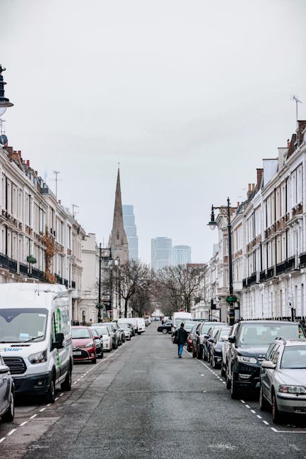 A narrow residential street in Golborne Road, Ladbroke Grove, with a row of terraced houses on each side. The houses are a mix of white and brick facades, with some featuring small front gardens filled with potted plants and trees. The street is paved with cobblestones and has a white van parked near the center, likely used for a home relocation or furniture transport. Several large cardboard boxes and wrapped pieces of furniture are visible, being loaded or unloaded by movers, who are partly visible near the van. Tools such as trolleys and straps may be seen in use during the loading process. The scene is under a clear, daytime sky with modern buildings visible in the distance, highlighting the typical environment for house removals and packing during moving services provided by Man With a Van Ladbroke Grove.