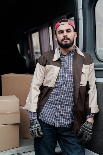 A man wearing a white t-shirt and jeans is stacking cardboard boxes on the pavement beside a white moving van with its rear doors open. The boxes vary in size and are arranged in a neat sequence, with
