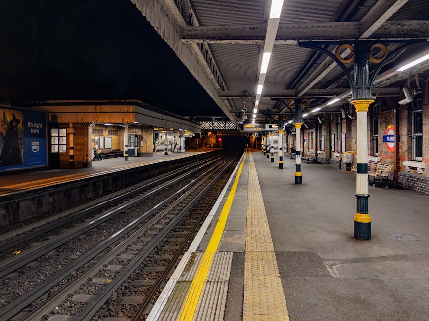 A view of an empty underground railway station platform at night, with the tracks running parallel along the centre. The platform is bordered by a yellow safety line and tactile paving for visually impaired passengers. The station features brick walls, some with advertising posters, and metal columns with decorative yellow and black bands supporting the ceiling. In the distance, a few benches are visible, and a small station building is illuminated on the far side. The scene is well-lit with overhead lighting, emphasizing the empty space and quiet atmosphere. This environment is suitable for a home relocation process, where furniture and boxes might be loaded onto nearby vehicles, such as a van from Man With a Van Ladbroke Grove, which could be involved in transporting household items from this site.