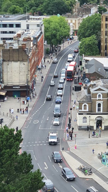 An aerial view of a busy street near Ladbroke Grove station showing various vehicles including cars, vans, and a double-decker bus moving along the road. On the left side, there are red brick and grey commercial buildings with ground-floor shops and pedestrians walking on the sidewalk. The right side features residential-style buildings with small storefronts and bicycles parked near the pavement. Several large, leafy green trees line both sides of the street, providing shade and greenery to the urban environment. The scene captures the typical movement and activity associated with home relocation and furniture transport processes, possibly related to a professional removals service like Man With a Van Ladbroke Grove, as they assist with packing, loading, and transporting household items through this area. The lighting suggests daytime with clear weather, highlighting the mix of commercial and residential structures in this lively neighbourhood.