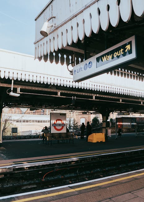 A view of an empty underground railway station platform at night, with the tracks running parallel along the centre. The platform is bordered by a yellow safety line and tactile paving for visually impaired passengers. The station features brick walls, some with advertising posters, and metal columns with decorative yellow and black bands supporting the ceiling. In the distance, a few benches are visible, and a small station building is illuminated on the far side. The scene is well-lit with overhead lighting, emphasizing the empty space and quiet atmosphere. This environment is suitable for a home relocation process, where furniture and boxes might be loaded onto nearby vehicles, such as a van from Man With a Van Ladbroke Grove, which could be involved in transporting household items from this site.