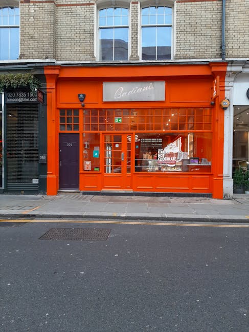 The image depicts the storefront of a retail shop with a bright orange facade, including large glass windows and a glass door, situated on a city pavement. The signage above the entrance reads 'Badiani' in elegant white script. Inside the window display, various boxed products and promotional items are visible, while the interior lighting illuminates the space. To the left of the shop, part of a neighboring premises with a dark-colored entrance is visible, and to the right, there is a glimpse of another store with a partially visible sign. The upper part of the building features beige brickwork and tall windows. The scene appears to be in a busy urban area, and the shop's vibrant exterior contrasts with the neutral tones of the surrounding buildings. Man With a Van Ladbroke Grove, a removals service, may use such storefronts or nearby properties during home relocation or furniture transport processes, as part of their house removals or packing and moving service offerings.