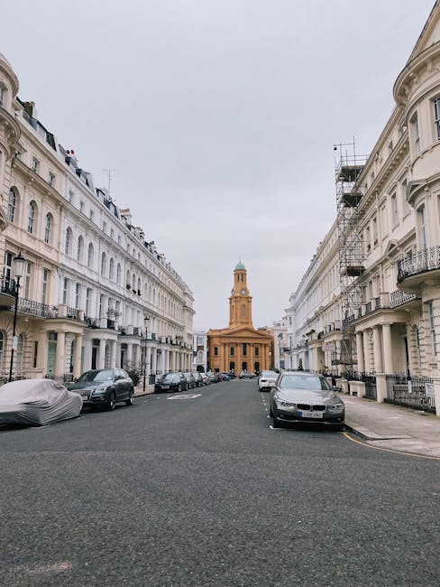 Photograph of a narrow residential street in Ladbroke Grove, with white multi-story Victorian-style buildings lining both sides. Parked cars are visible along the curbs, including a covered vehicle on the left and a modern grey car on the right. In the background, there is a prominent church with a tall clock tower featuring a green dome at the top, situated at the end of the street. The sky above is overcast, casting diffuse light over the scene. The image relates to local house removals and furniture transport, illustrating an urban environment where packing and loading processes could take place as part of a home relocation project by Man With a Van Ladbroke Grove.
