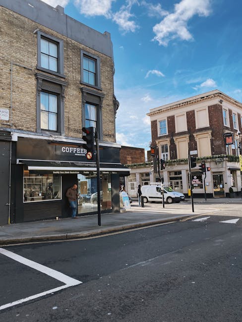 The image depicts the storefront of a retail shop with a bright orange facade, including large glass windows and a glass door, situated on a city pavement. The signage above the entrance reads 'Badiani' in elegant white script. Inside the window display, various boxed products and promotional items are visible, while the interior lighting illuminates the space. To the left of the shop, part of a neighboring premises with a dark-colored entrance is visible, and to the right, there is a glimpse of another store with a partially visible sign. The upper part of the building features beige brickwork and tall windows. The scene appears to be in a busy urban area, and the shop's vibrant exterior contrasts with the neutral tones of the surrounding buildings. Man With a Van Ladbroke Grove, a removals service, may use such storefronts or nearby properties during home relocation or furniture transport processes, as part of their house removals or packing and moving service offerings.